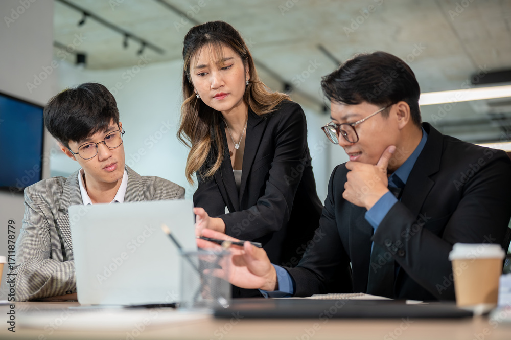 Φωτογραφία Asian businesswoman frowning while looking at laptop aside male coworkers on wooden table in office