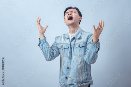 Asian man in denim jacket screaming with both hands raised, expressing anger, frustration, sadness, or despair on a plain studio background, dramatic emotional reaction