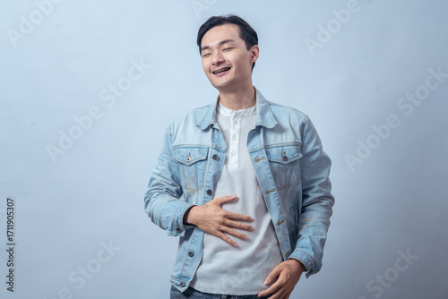 Asian man in denim jacket touching stomach with pained face, standing in studio against plain light background, expressing stomachache, discomfort, and sick feeling with casual fashion style
