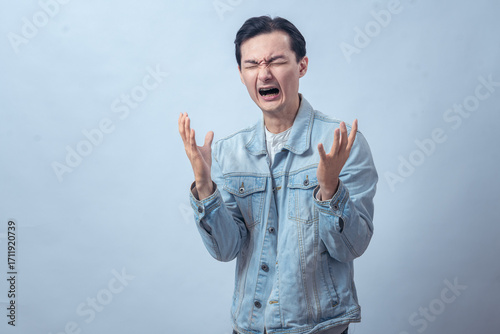 Asian man in denim jacket screaming with both hands raised, expressing anger, frustration, sadness, or despair on a plain studio background, dramatic emotional reaction