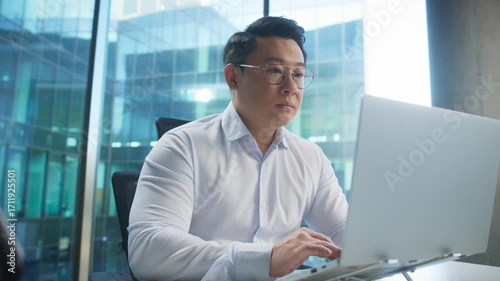 Determined Asian man in formal shirt wearing glasses and typing on laptop in modern workspace. Responding to urgent email or preparing business proposal. Working before deadline on report.