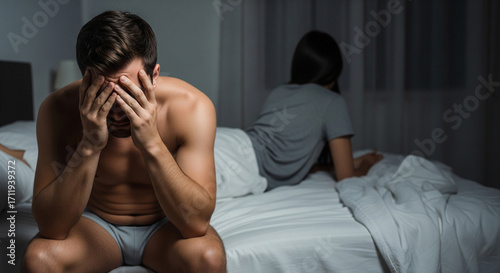 Man disappointed in himself due to premature ejaculation and erection problems, upset sitting on bed holding head, lost self-confidence due to dissatisfied and disappointed wife lying on bed 