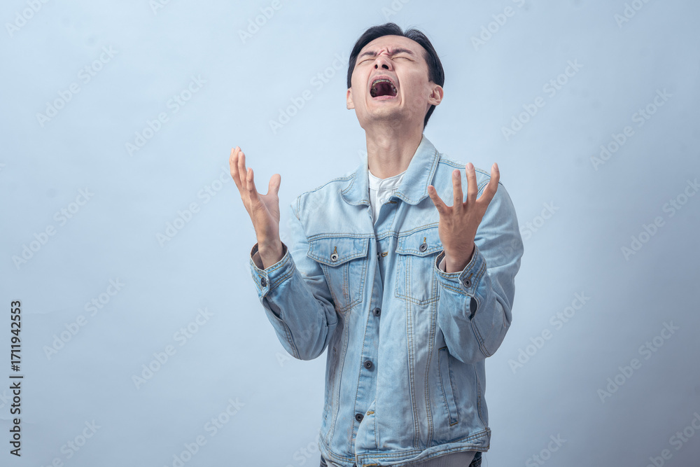 Naklejka premium Asian man in denim jacket screaming with both hands raised, expressing anger, frustration, sadness, or despair on a plain studio background, dramatic emotional reaction