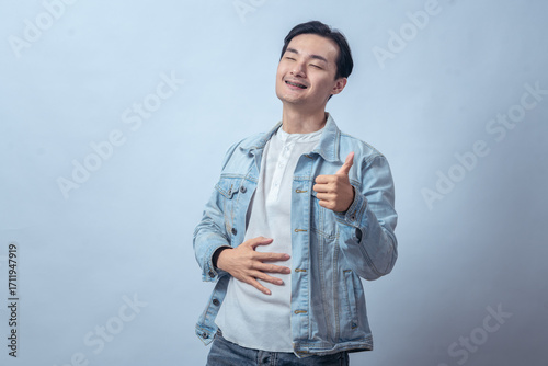 Asian man in denim jacket smiling with eyes closed, touching stomach with one hand and showing thumbs up, standing in studio against plain light background, expressing relief and satisfaction