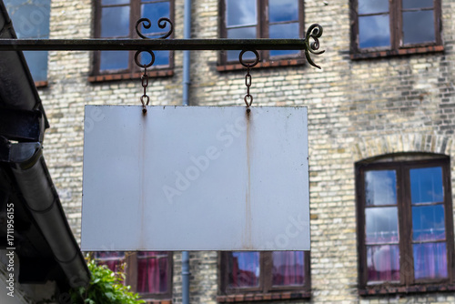 Blank Rectangular White Sign Hangs From A Wrought-iron Arm In Front Of A Weathered Brick Building