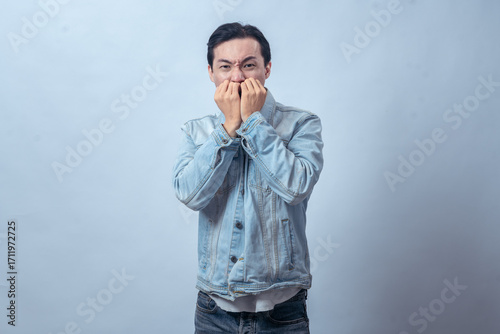 Asian man in denim jacket biting nails with anxious face and tense body language, standing in studio against plain light background, expressing nervousness, stress, and fear