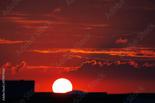 Stunning fiery sunset scene with vibrant red sky glowing sun setting behind city buildings urban silhouettes, creating warm dramatic atmosphere.  Tranquil horizon dusk cloudscape