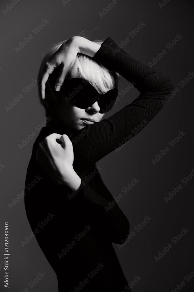 Obraz premium Stylish woman in black outfit striking a pose with bold sunglasses, showcasing confidence and fashion in a dramatic black and white setting.