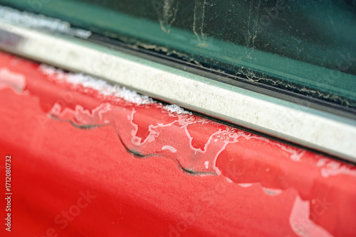 Damaged red car paint on door with peeling clear coat and faded paintwork, oxidation, deterioration, and visible surface wear on the exterior finish of old vehicle. Selective focus