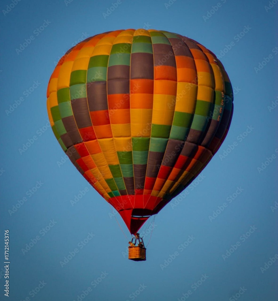 Naklejka premium A colorful hot air balloon floats in a clear blue sky. The balloon features a vibrant pattern of orange, green, and yellow panels. It is a serene scene of flight.