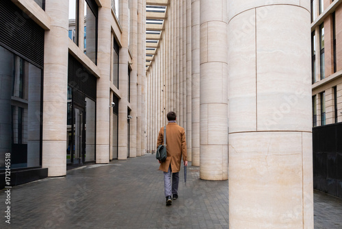 man in autumn coat walks under colonnade in business district of city, down town. man in full growth walks away from the camera. businessman, lawyer, manager, office worker