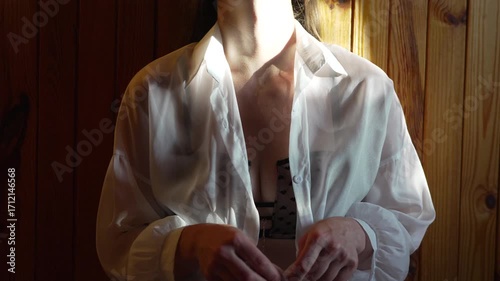 Woman undressing white shirt, revealing black bra in sunlit wooden room