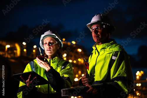 Team of construction engineers in uniforms and helmets examine project with tablet Industrial plant or oil refinery background at night