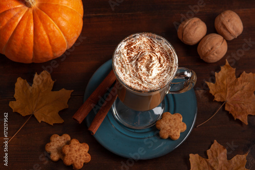 A top-down view of a pumpkin spice latte with whipped cream and cinnamon powder, surrounded by a pumpkin, nuts, leaves and cookies.