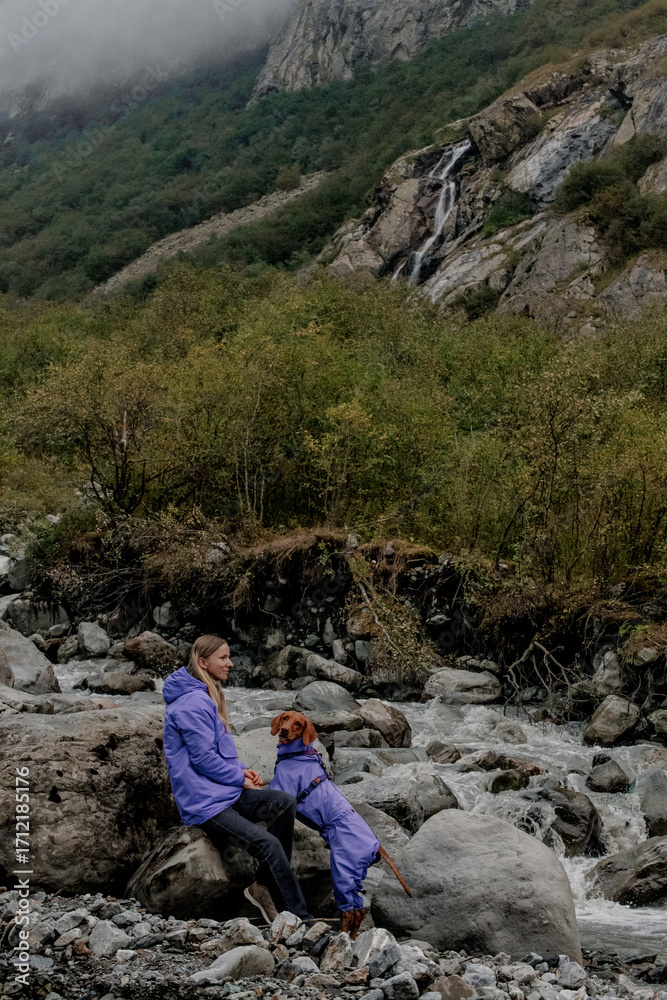 Young Caucasian man and a child sit on rocks by a river in a mountainous area. Both wear purple jackets. Lush greenery surrounds the scene. Young Caucasian man and a child sit on rocks by a river in a mountainous area. Both wear purple jackets. Lush greenery surrounds the scene.
