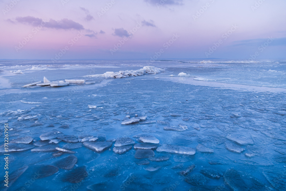 Fototapeta premium Frozen ocean with cracked ice and stacked slabs under a pastel pink and blue sunset sky in winter stillness.