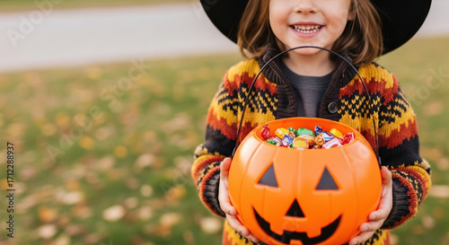 Little girl trick or treating on Halloween, holding a bright pumpkin basket filled with candy. Halloween celebration finds toddler in costume with treats.