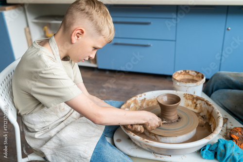 Blond boy learning pottery, close-up of hands shaping clay on a wheel, creative kids activity in cozy pottery studio, teamwork and manual craft skills concept