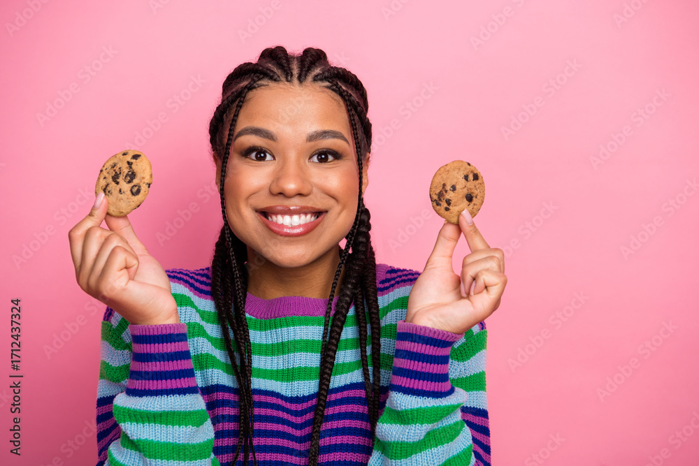Naklejka premium Cheerful young woman in a colorful outfit holds chocolate chip cookies against a pink background with a bright smile