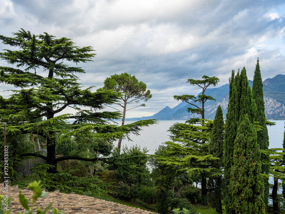Fototapeta premium Ancient castle walls rise above Lake Garda, surrounded by tall cypress trees and lush greenery. The view opens to dramatic cliffs and blue water under a cloudy sky in northern Italy.