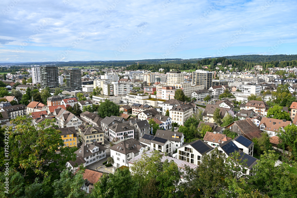Naklejka premium Panoramic view of Uster, Switzerland, showing the church tower with clock, residential rooftops, modern buildings, and green surroundings under a partly cloudy sky
