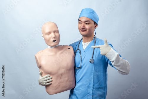 Asian doctor in blue scrubs smiling with braces, holding a CPR training mannequin and showing an OK gesture, wearing gloves and stethoscope against plain background
