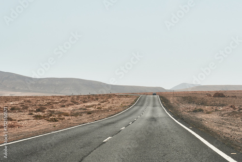 Photograph of a solitary asphalt road across the dry desert landscape of Fuerteventura, with low mountains in the distance.
