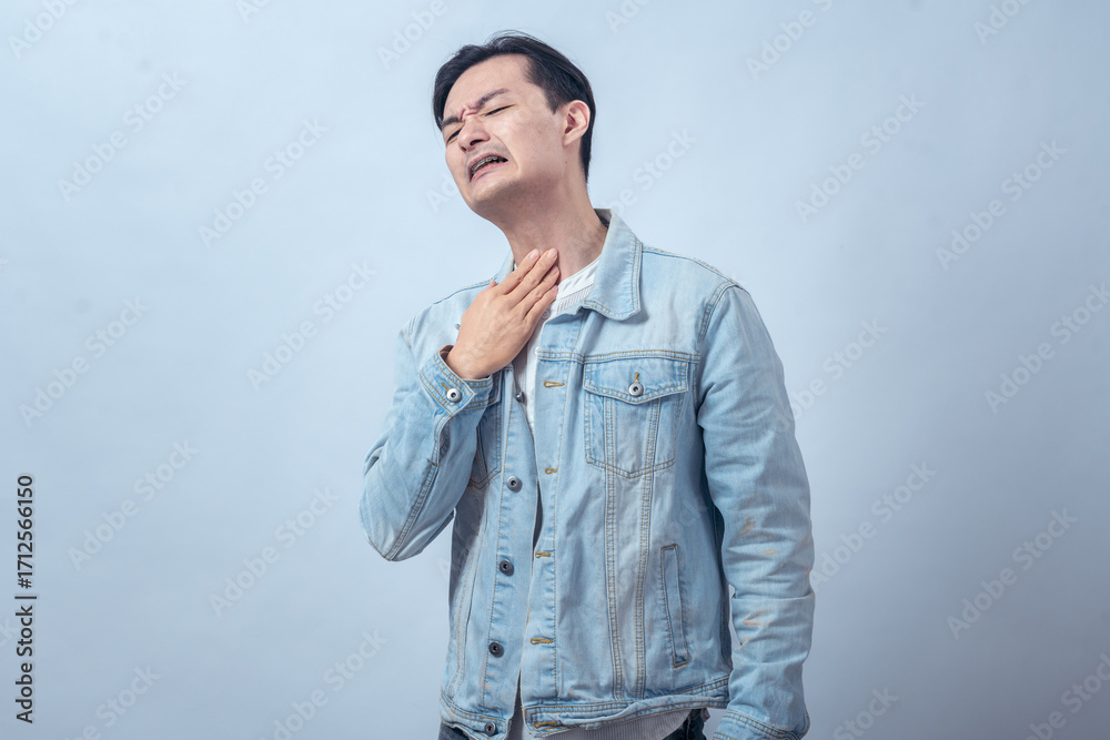 Fototapeta premium Asian man in denim jacket clutching his throat with distressed expression, standing in studio against plain light background, showing choking, breathing difficulty, and pain