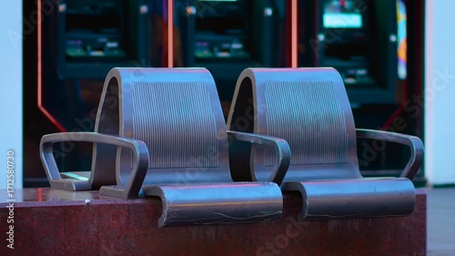 Chairs placed in front of brightly lit ATMs during evening time. Loungers standing still near cash dispensers on quiet urban street. Seats appearing vacant by glowing machines in city twilight