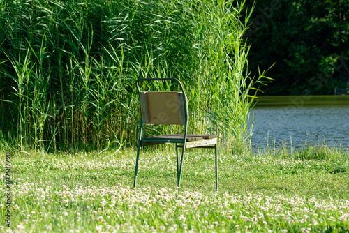 An old chair sits peacefully on the green grass near a lake on a sunny day.