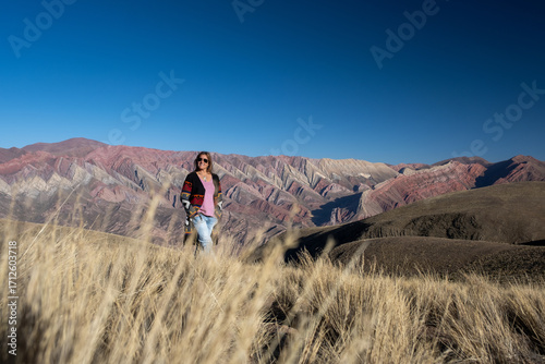 A smiling woman stands in golden grass, admiring the breathtaking, multi-colored Serranía del Hornocal mountains in Jujuy, Argentina. A vibrant travel adventure