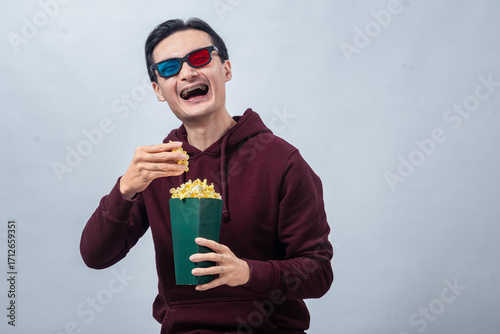 Asian man in maroon hoodie wearing 3D glasses, happily eating popcorn from a green container while seated against a plain background