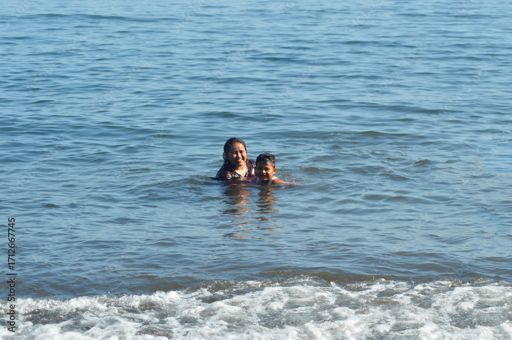 Fototapeta premium A cheerful mother walks toward her young son in the shallow surf, smiling warmly as they enjoy playful water activities together in the gentle morning tide at the beach.