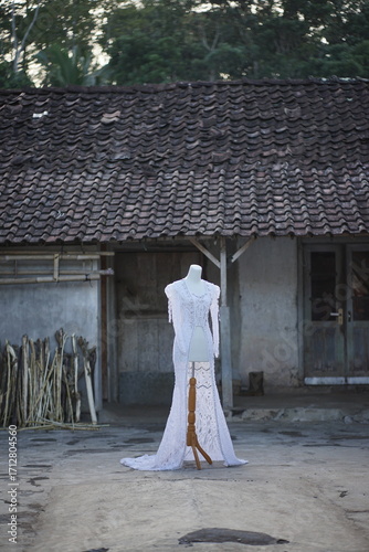 Elegant white bridal gown on a classic mannequin, displayed in a tranquil rustic outdoor setting with an aged building, embodying timeless fashion and enduring design