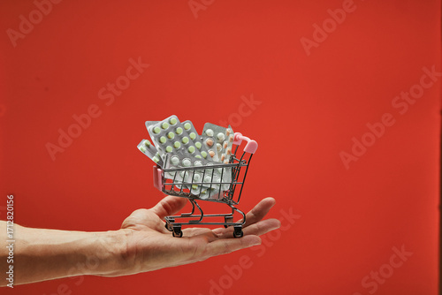 Hand gripping tiny shopping cart containing medication blister packs against vibrant red surface, symbolizing escalating pharmaceutical expenses