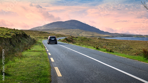 Car driving on scenic country road in Connemara Ireland with mountains and lake at colorful sunset sky, nature background