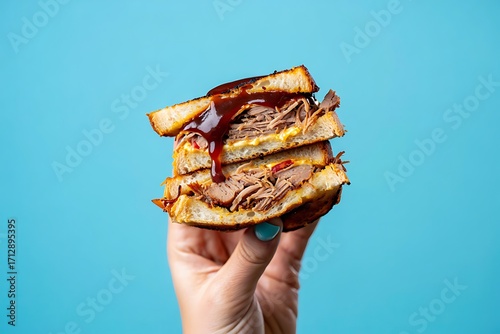 Hand holding a delicious pulled pork sandwich stacked high against a blue background