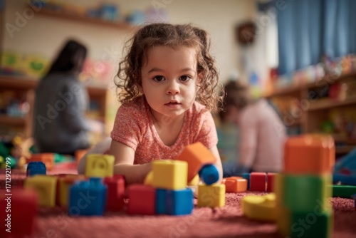 Young girl playing with colorful toy blocks in a bright child care center while adults engage in activities in the background