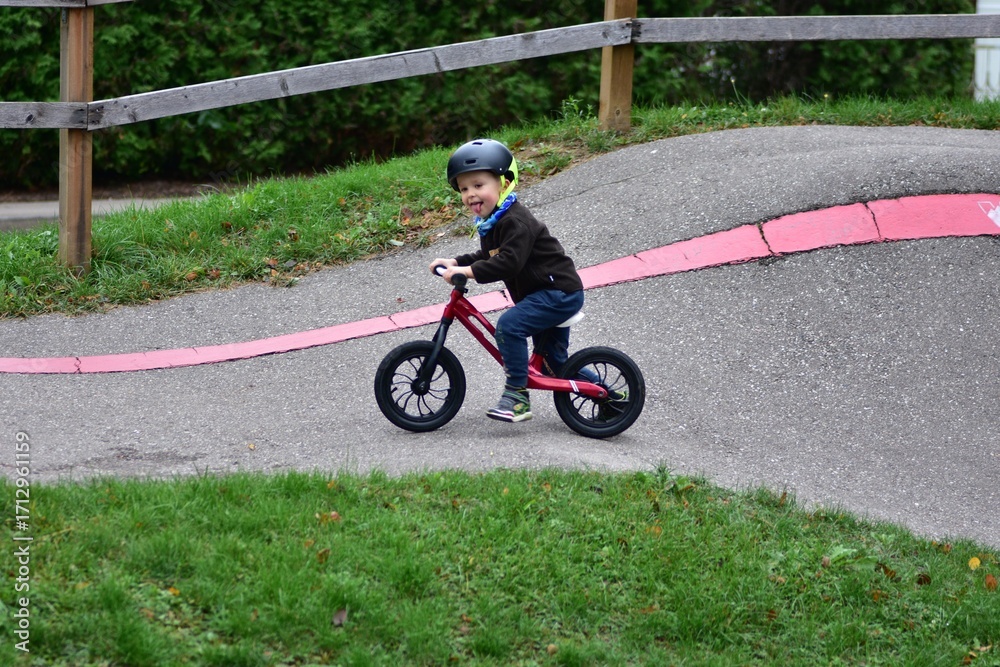 Obraz premium Young boy riding balance bike with helmet on curved asphalt track. Child enjoys sport activity, balancing carefully while cycling outdoors on pump track in autumn season.