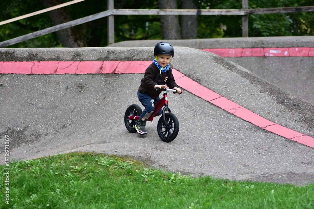 Obraz premium Young boy riding balance bike with helmet on curved asphalt track. Child enjoys sport activity, balancing carefully while cycling outdoors on pump track in autumn season.