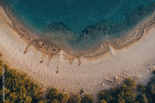 Fototapeta Naklejka Na Ścianę i Meble -  Overhead perspective of a sandy spit protected by a barrier at dusk. Aerial shot of a white beach meeting clear blue waters. Coastal scenery with transparent turquoise sea. Natural landscape seen