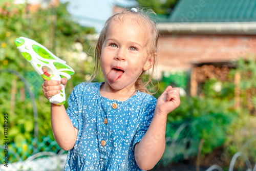 A little child girl, wet with water, runs from a watering can with a water pistol in her hands in summer