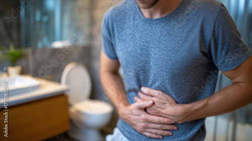 Side profile of person with clasped hands over stomach, open toilet behind, bathroom interior with muted colors, sense of discomfort and urgency conveyed