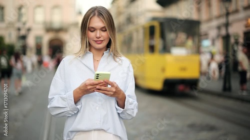 Young woman walks along a city street, holding smartphone and texting or scrolling social media feed. Yellow tram passes in the background, creating an urban lifestyle atmosphere
