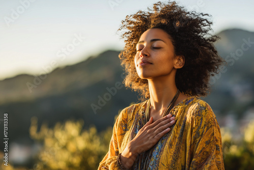 Woman with curly black hair and brown sweater holding hands over heart in lush green outdoor setting with blurred foliage