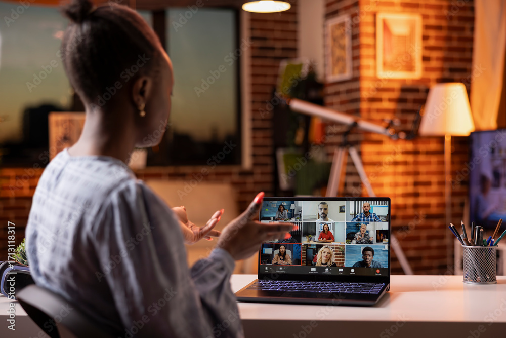 Fototapeta premium Black woman joining video call with team, working from home on laptop after sunset. Focused female entrepreneur in virtual meeting, handling evening business discussions with remote colleagues.