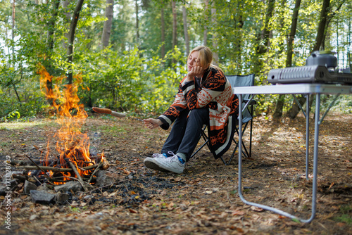 Young woman relaxing by campfire and grilling sausages in forest