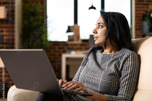 Fotografie Indian businesswoman sits on sofa, looking towards window with pensive expression