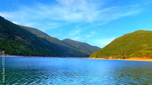 Heavenly scene from Lago di Fiastra in the Sibillini Mountains, the fluid mirroring rippling waters of the lake surrounded by shores, peaks with vegetation and a dam, under a blue sky with thin clouds