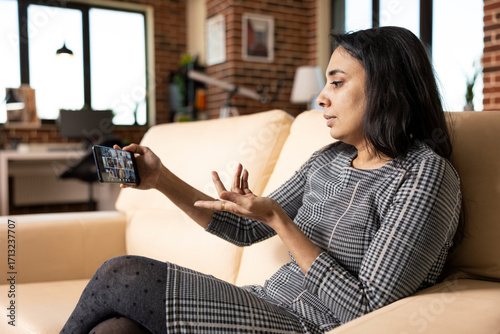 Canvas Print Indian female executive seated on sofa with cellphone, speaking remotely with diverse employees on video call